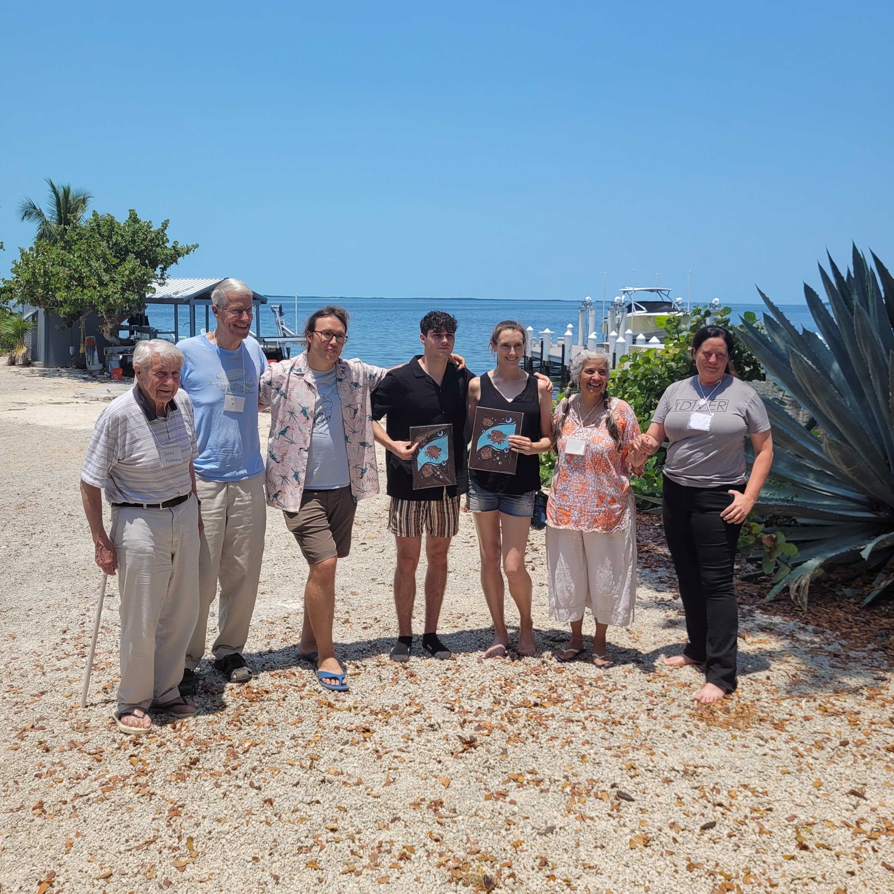 Group of people on beach with water in background, two holding awards